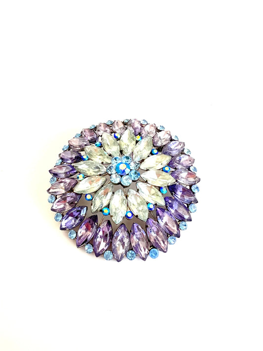 A round floral brooch featuring faceted rhinestones in shades of purple, lavender, clear, and light blue, set in a silver-tone metal on a white background.