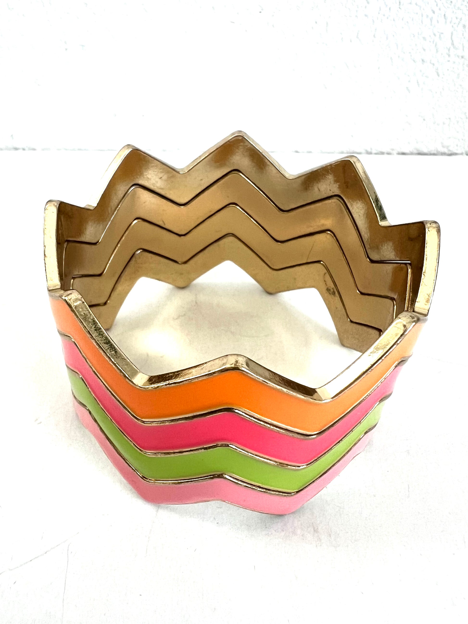 A chevron-style cuff bracelet with multiple stacked zigzag bands in gold, orange, pink, and light green, photographed on a plain white background.