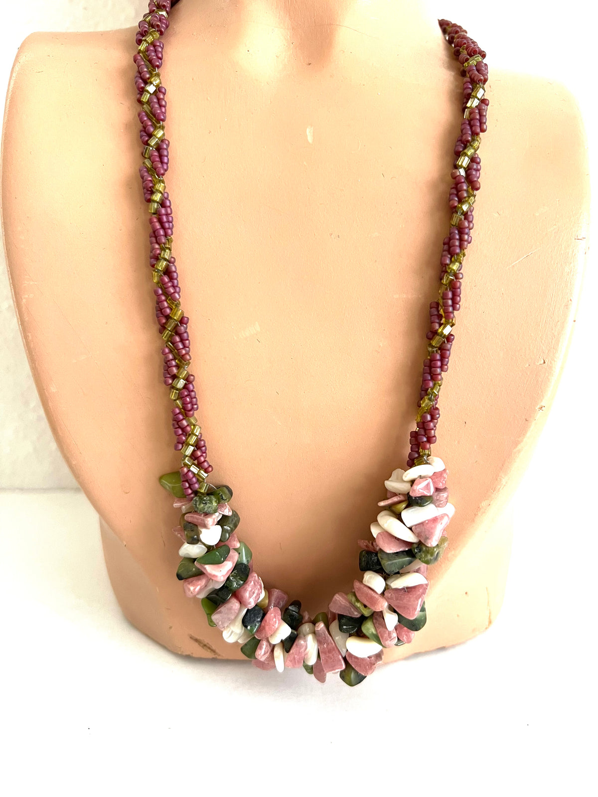 A handmade statement necklace displayed on a beige mannequin bust. The necklace features a braided section of purple and olive green beads, transitioning into a large cluster of polished pink, white, and green stone chips.