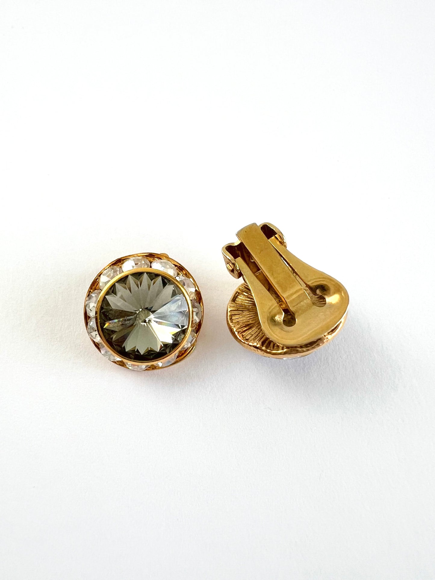 A pair of gold-toned round clip-on earrings, one showing the front with a faceted clear crystal and a halo of small clear stones, and the other showing the gold-tone clasp mechanism, on a white background.