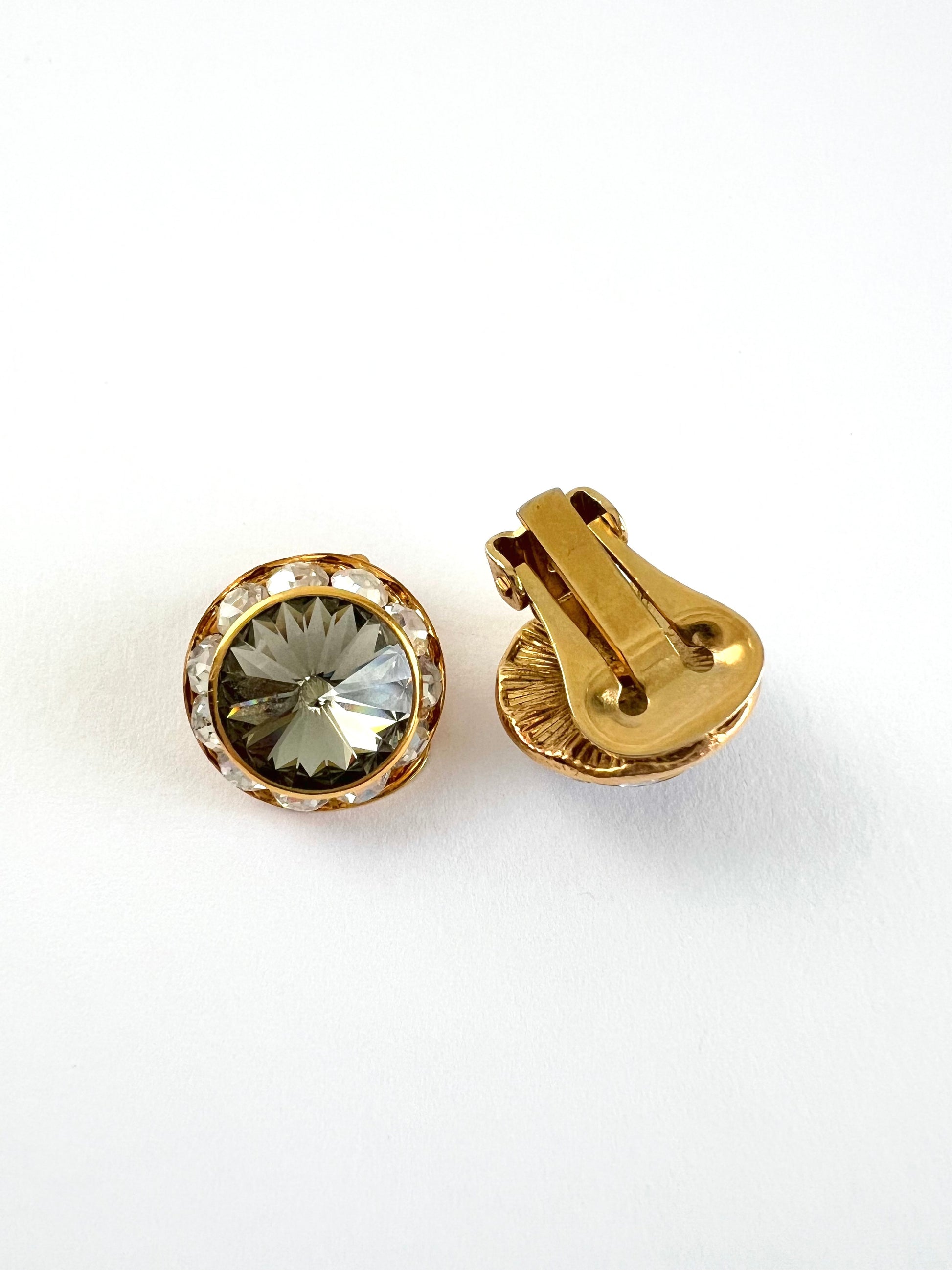 A pair of gold-toned round clip-on earrings, one showing the front with a faceted clear crystal and a halo of small clear stones, and the other showing the gold-tone clasp mechanism, on a white background.
