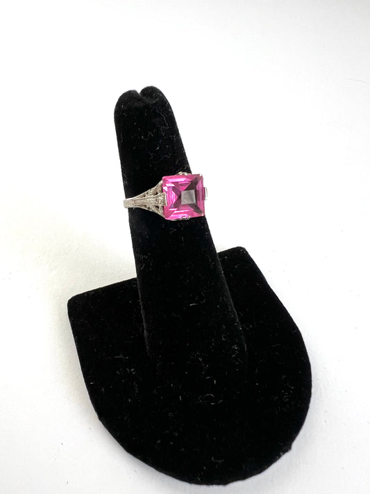 A silver ring with a square-cut pink gemstone displayed on a black velvet ring stand against a white background.