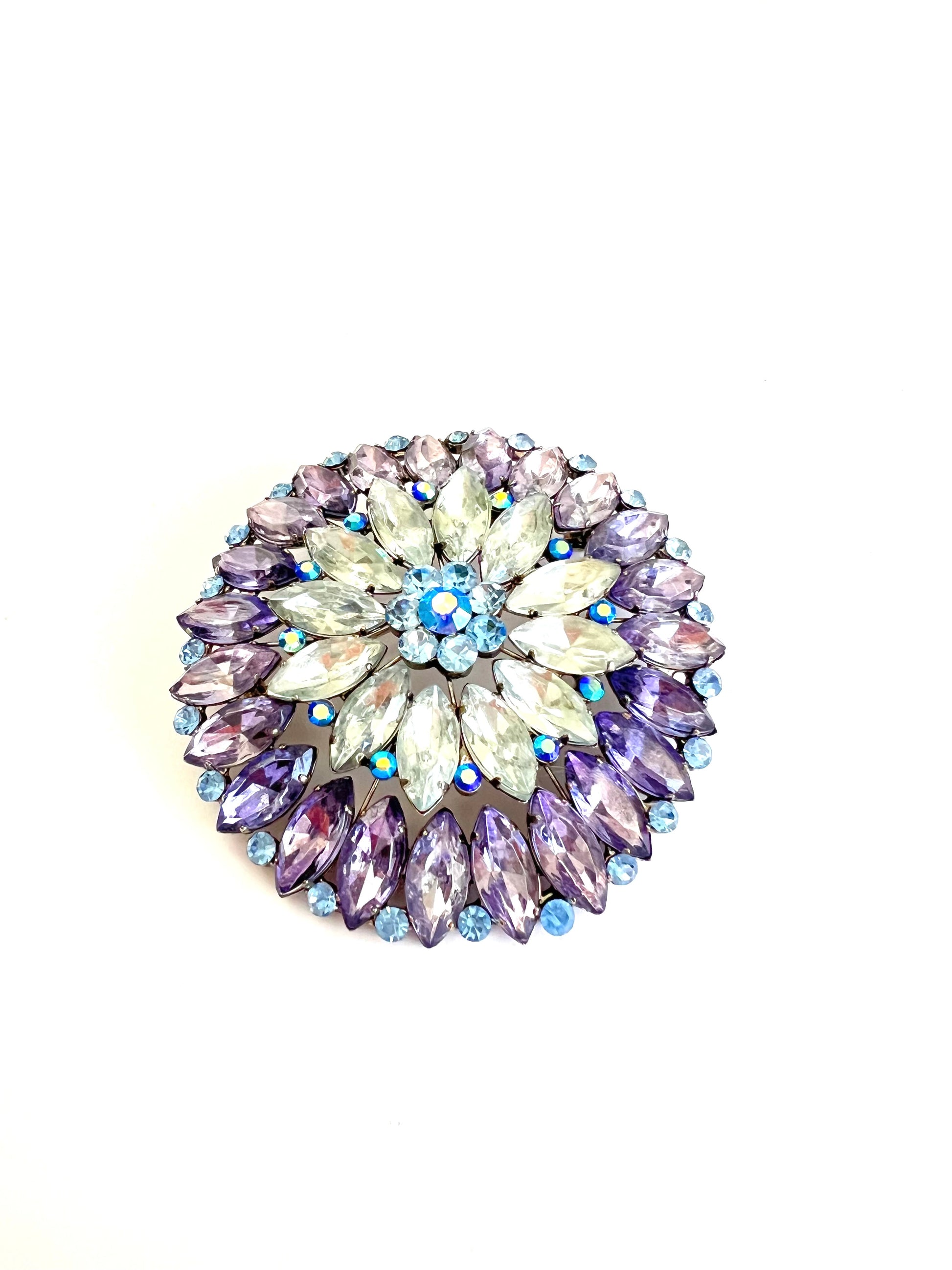 A round floral brooch featuring faceted rhinestones in shades of purple, lavender, clear, and light blue, set in a silver-tone metal on a white background.