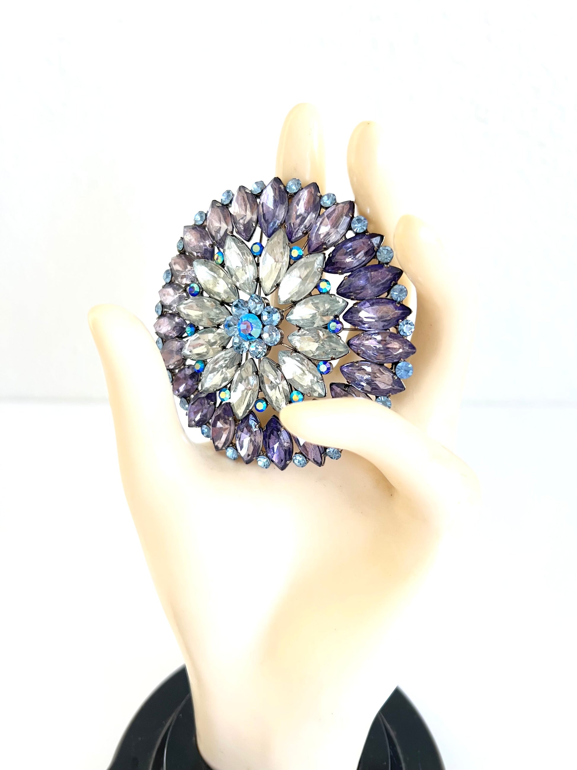 A large circular floral brooch with faceted purple and clear rhinestones, held in a cream-colored mannequin hand against a plain white background.