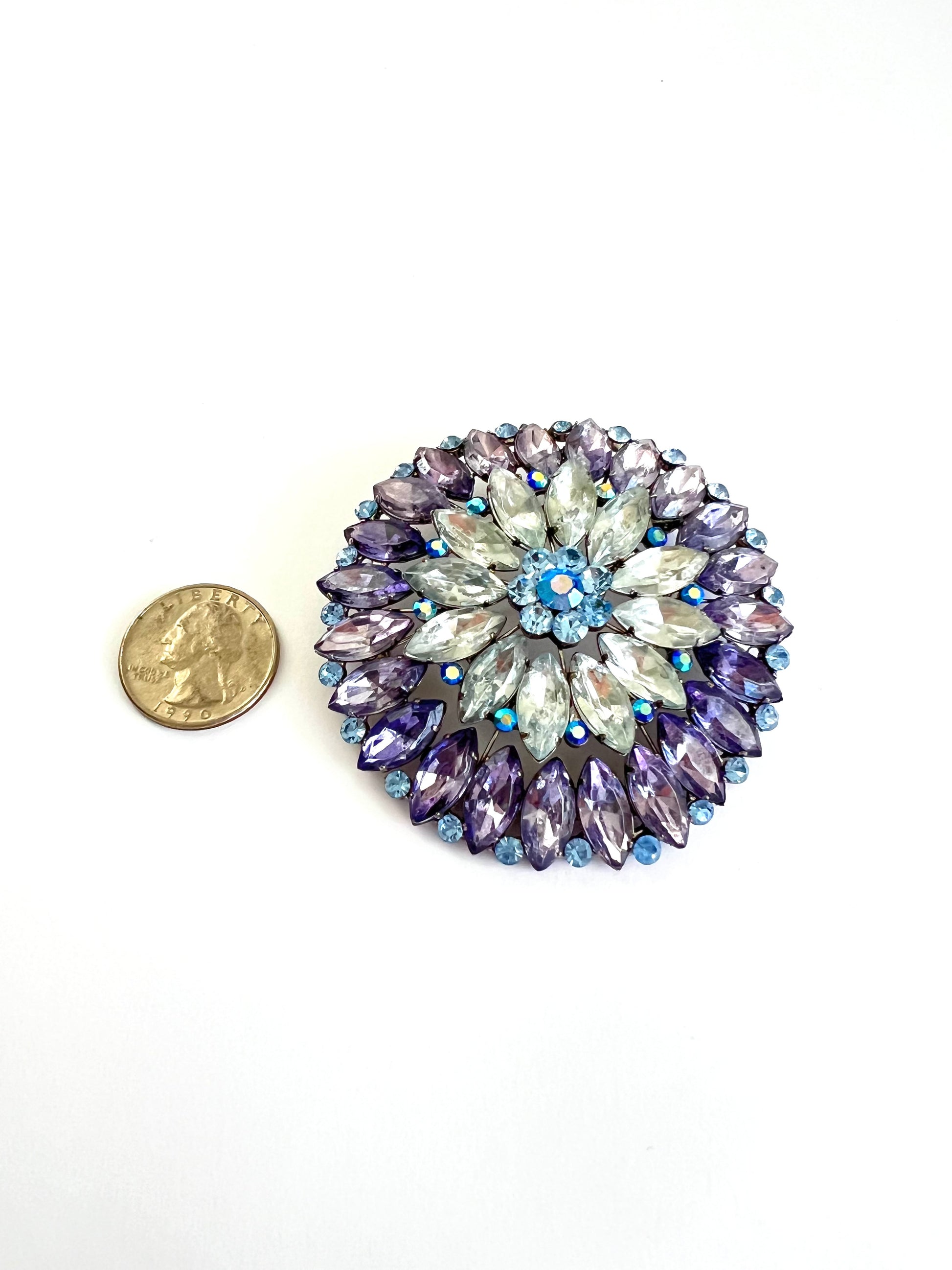 A round floral brooch with faceted purple, lavender, and light blue rhinestones, shown next to a US quarter coin for scale on a white background.