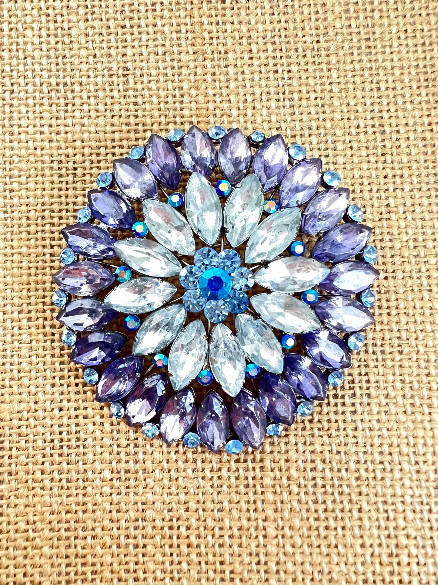 A circular floral brooch featuring light blue marquise-cut rhinestones in the center and purple marquise-cut rhinestones around the border, accented with small blue iridescent rhinestones, displayed on a beige burlap background.
