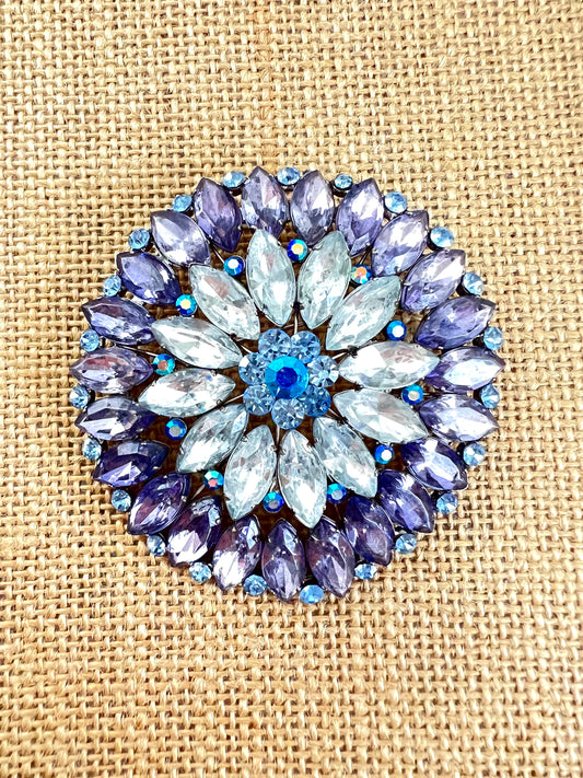 A circular floral brooch featuring light blue marquise-cut rhinestones in the center and purple marquise-cut rhinestones around the border, accented with small blue iridescent rhinestones, displayed on a beige burlap background.