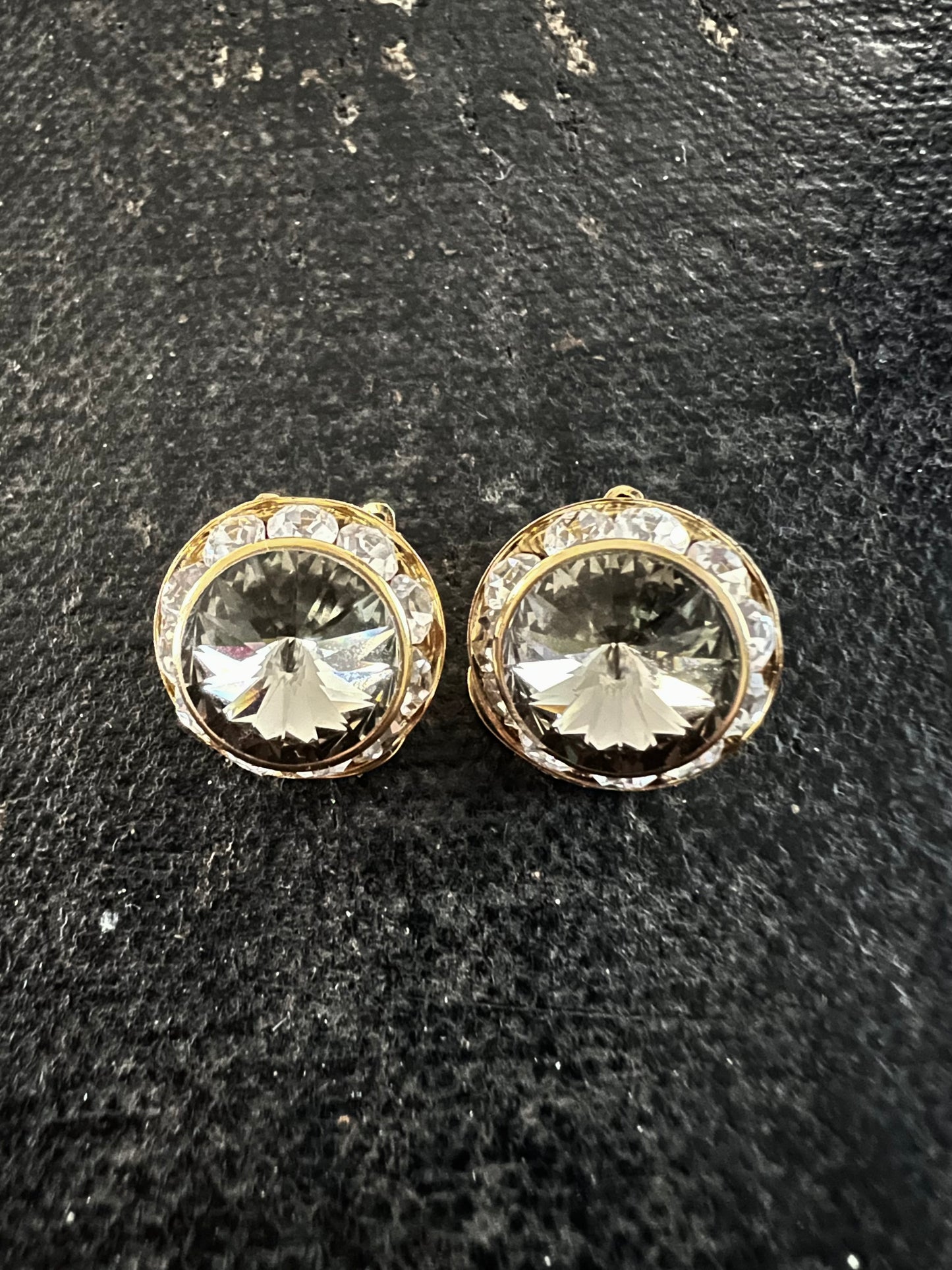 A pair of round gold-tone stud earrings with clear faceted center stones and a halo of small clear stones, displayed on a dark textured surface.