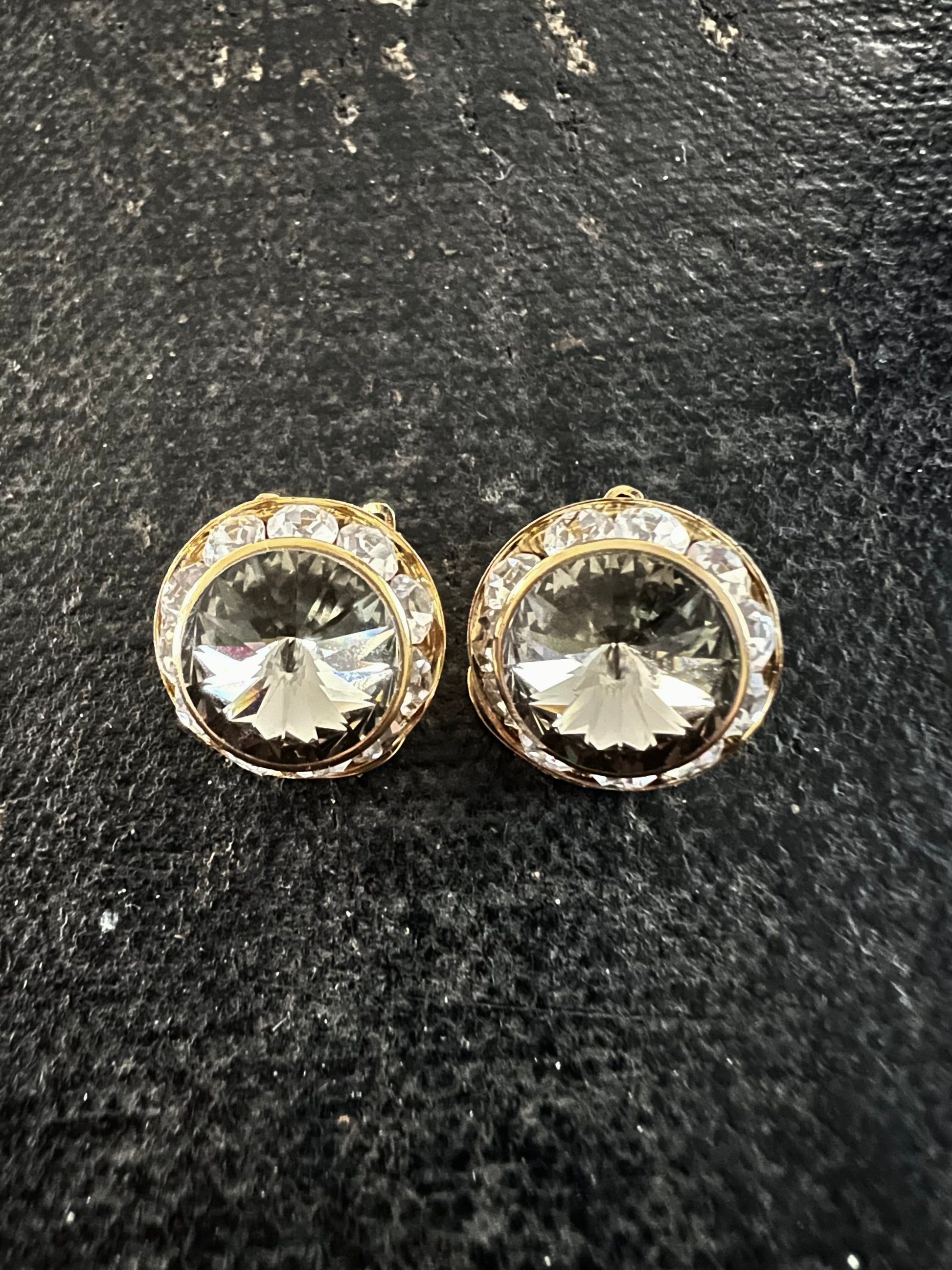 A pair of round gold-tone stud earrings with clear faceted center stones and a halo of small clear stones, displayed on a dark textured surface.
