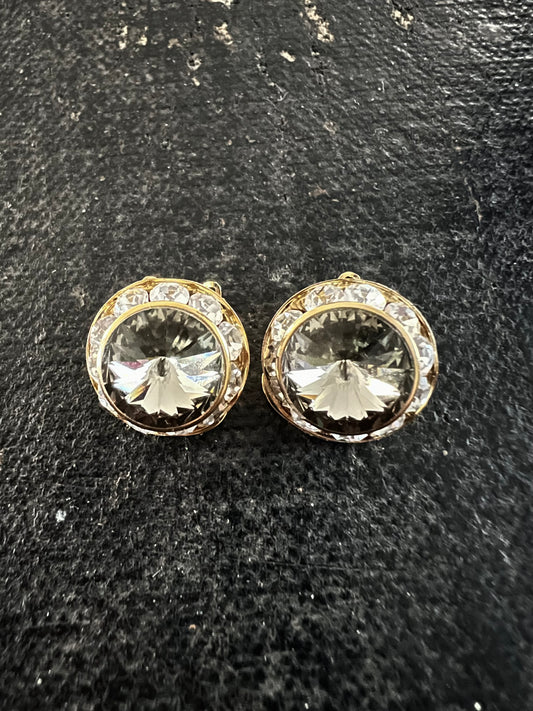 A pair of round gold-tone stud earrings with clear faceted center stones and a halo of small clear stones, displayed on a dark textured surface.
